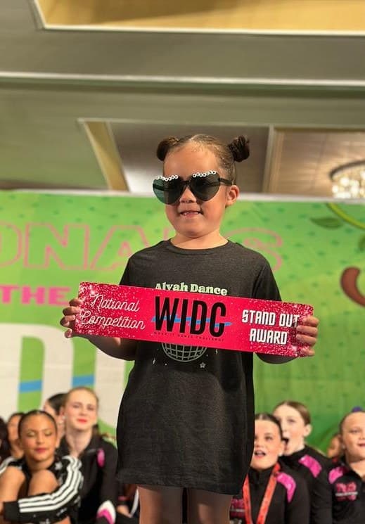 Young dancer in heart-shaped sunglasses holding a WIDC Stand Out Award sash at Nationals